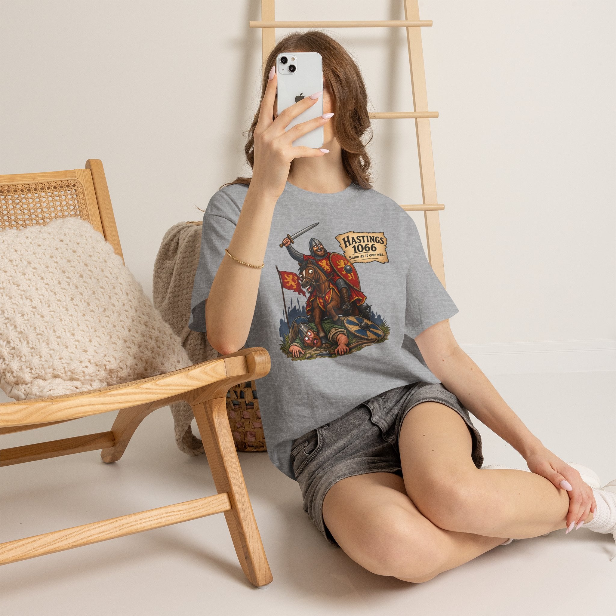 Person wearing a gray Battle of Hastings 1066 T-shirt with a graphic design, sitting on the floor in a room with a wooden chair and ladder.