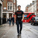 A man walking on a London street wearing a black graphic t-shirt with a red and white text design that reads 'BLACK DEATH ALLERGIC TO BATS SINCE 1987' alongside a bat silhouette. The background features classic London architecture, a red double-decker bus, and a black cab.