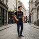 A man stands on a cobblestone street wearing a black graphic T-shirt with red and white text, dark jeans, and black shoes, while carrying a brown guitar. The background shows a narrow city street with buildings, shopfronts, and a red double-decker bus.