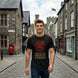 A man standing on a cobblestone street wearing a black t-shirt with a red dragon graphic and the text 'BORN OF FIRE MADE OF LEGEND'. The background shows a stone town street with a castle in the distance.