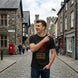 A man standing on a cobblestone street wearing a black graphic t-shirt with a red circular emblem and the text 'BORN OF FIRE MADE OF LEGEND' in gold. He is pointing to his chest, with stone buildings and a castle in the background.