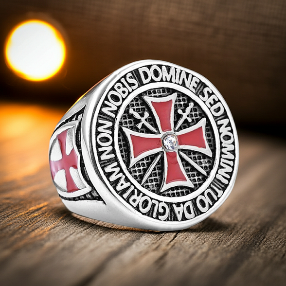 A silver-toned ring featuring a red and black Templar cross emblem with a small clear stone at the center, resting on a wooden surface with a warm, blurred background.