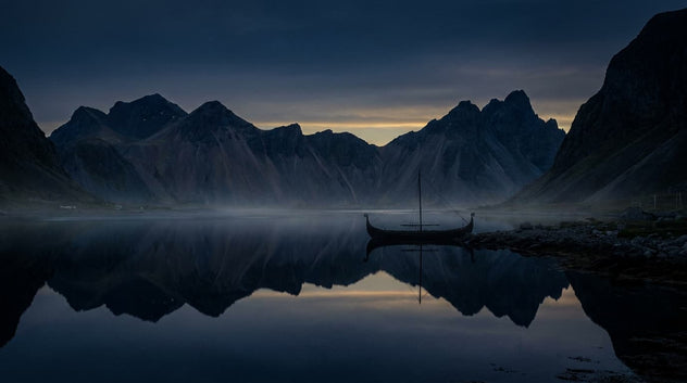 A dramatic mountain landscape at dusk or dawn with jagged peaks reflected in a calm lake, a small boat near the shore, and a dark blue sky above.