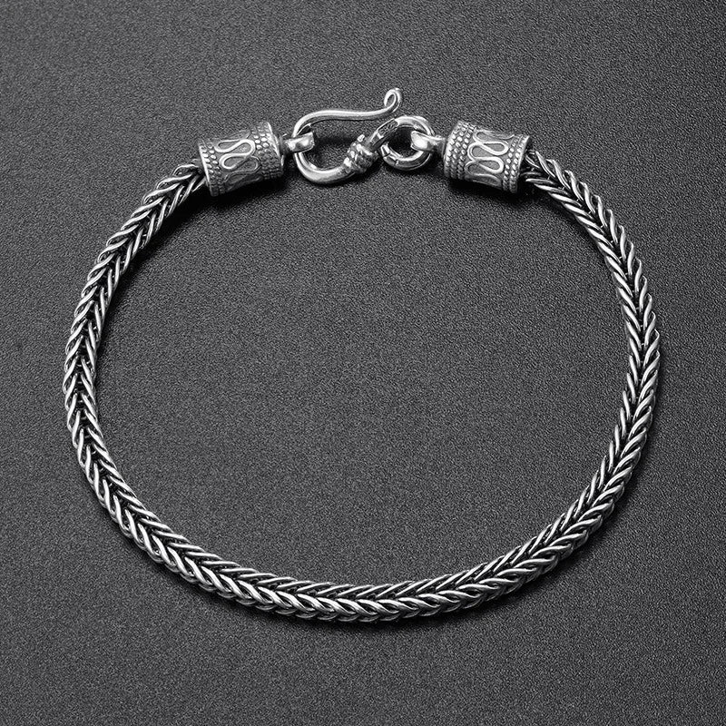 A silver-toned braided rope bracelet with a lobster clasp and decorative cylindrical end caps, photographed on a dark textured background.