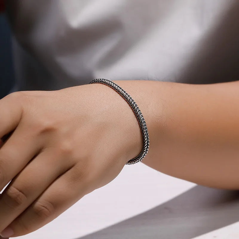 A close-up of a person's wrist wearing a thin braided metallic bracelet in a silver-tone finish, with a soft gray shirt blurred in the background.
