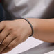 A close-up of a person's wrist wearing a thin braided metallic bracelet in a silver-tone finish, with a soft gray shirt blurred in the background.
