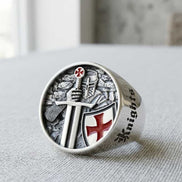 Silver ring with a detailed design featuring a knight, sword, and shield on a light fabric background.