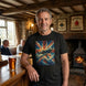 A man wearing a black graphic t-shirt stands in a rustic pub. The t-shirt features a colorful print of a vintage fighter plane flying against a Union Jack flag background.