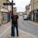 A man stands on a street corner wearing a black graphic t-shirt with a red dragon design and the text 'THE RED DRAGON RISES', paired with blue jeans. The background shows a real-world street scene with shops, a street sign, and a van.