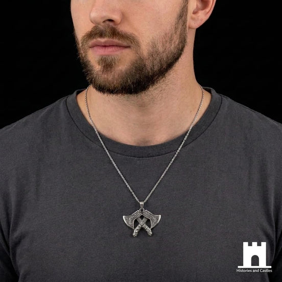 A close-up of a man wearing a silver necklace with a double-headed axe pendant, styled with a dark gray t-shirt against a black background.