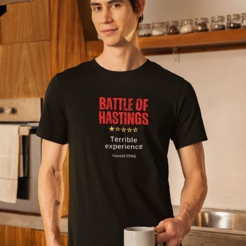 A man in a kitchen wears a black Battle of Hastings T-shirt featuring five stars with only one highlighted, Terrible experience, and Harold 1066. He holds a white mug and smiles slightly.