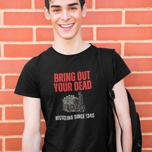 A smiling young person with dark hair and a backpack stands before a brick wall, wearing the “Bring Out Your Dead” – Medieval Plague T-Shirt featuring BRING OUT YOUR DEAD RECYCLING SINCE 1349 and a medieval cart graphic.