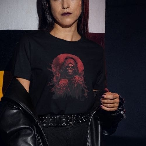 A woman in dark clothing and lipstick stands against a painted wall, wearing the Gothic Grim Reaper Floral T-Shirt – Beautiful for Dark Souls, featuring a hooded reaper with flowers. She holds a black leather jacket draped over her arm.