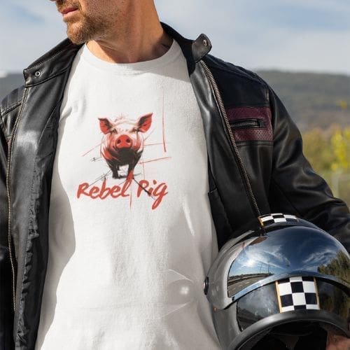 A man in a black leather jacket holds a checkered helmet and wears the Rebel Pig T-shirt, which showcases a stylized pig graphic and bold red text. He stands against a scenic backdrop of trees and hills.