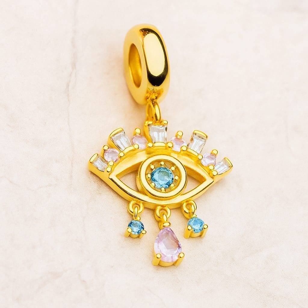 Gold eye-shaped pendant charm featuring a central Blue Nazar Eye gemstone, surrounded by pink and clear stones, with three hanging blue and pink gems, on a light textured background.
