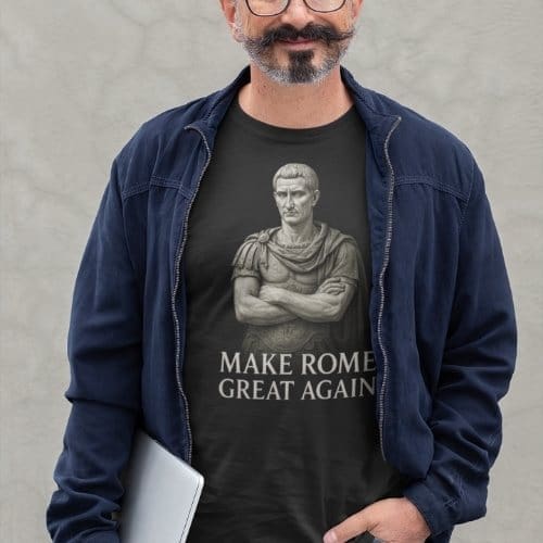 A man with glasses and a mustache, in a dark jacket and Make Rome Great Again T-shirt, featuring a Roman statue graphic, stands holding a closed laptop against a light gray background.