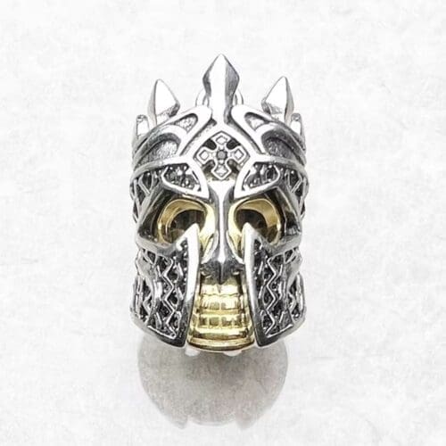 The Pendant Skull Knight is a detailed metal ring shaped like a skull in an ornate crown and helmet, with intricate silver patterns and golden accents on the face and teeth, shown against a white background.
