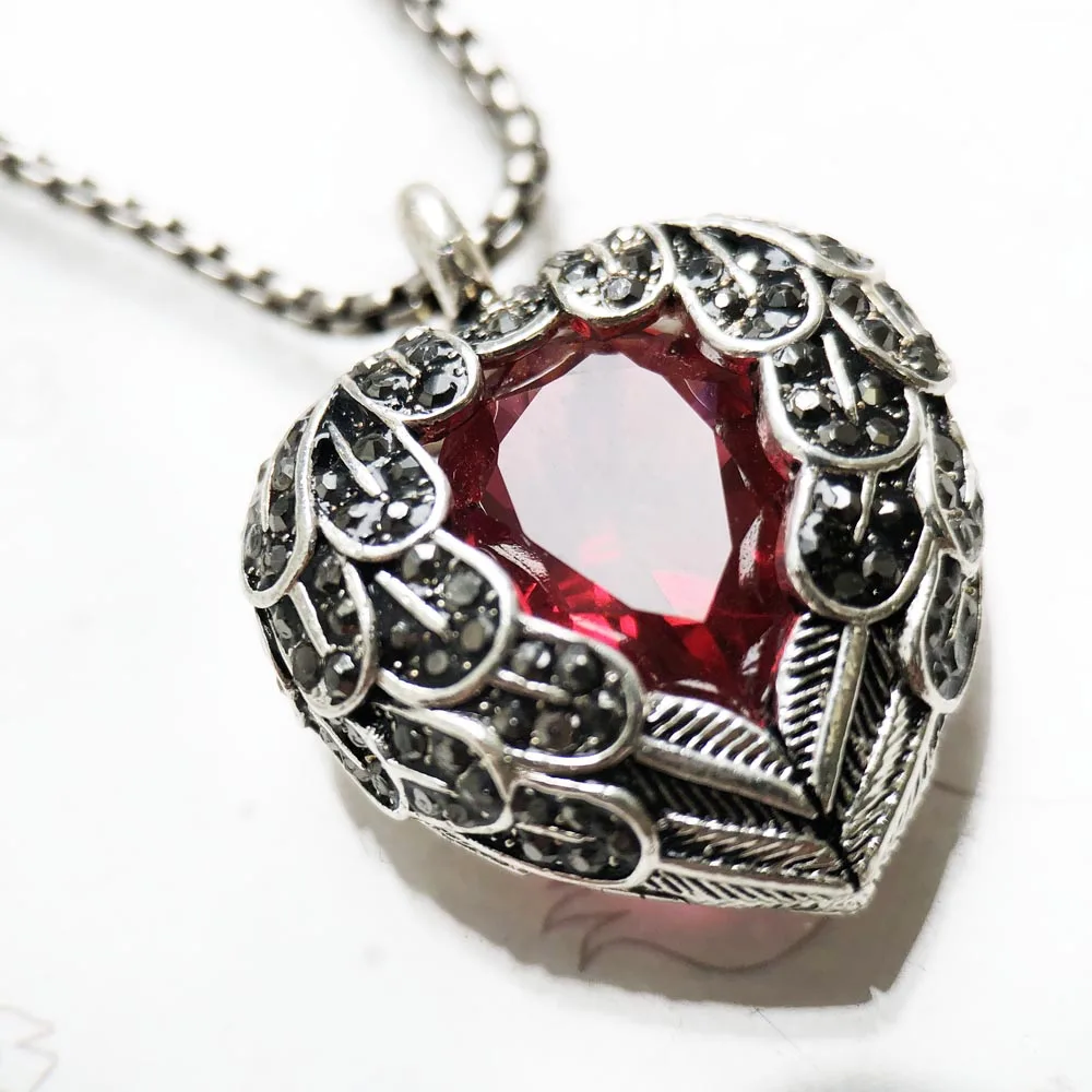 The Pendant Necklace Red Winged Heart showcases a large red winged heart gemstone in ornate silver with leaf and scale designs, attached to a silver chain.