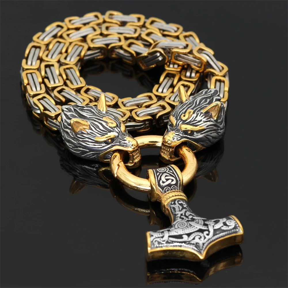 The Viking Wolf Necklace features two intricately detailed wolf heads holding a ring, from which a decorated Mjolnir pendant hangs, all set against a striking black background in gold and silver tones.