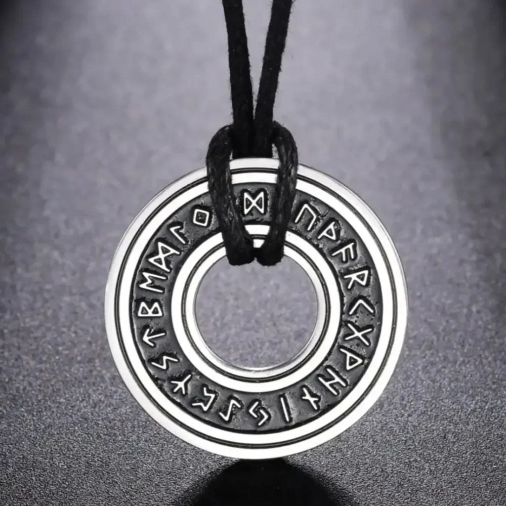 The Norse Runes Viking Pendant Amulet, a round metallic piece engraved with runes, hangs on a black cord and is shown against a dark, textured background.